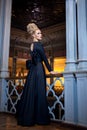 Gorgeous young blonde model, dressed in a long black dress with bow at the back, elegant hairstyle, crown and earrings. Royalty Free Stock Photo