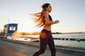 Gorgeous young girl with long brown hair dressed in sports clothes runs on the road along the reservoir on the sunset