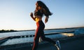 Gorgeous young girl with long brown hair dressed in sports clothes runs on the road along the reservoir on the sunset