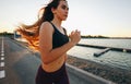 Gorgeous young girl with long brown hair in a sports top and tights runs on the road along the reservoir on the sunset