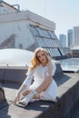 Gorgeous lady sitting on roof of high-rise building in white cotton dress and laced boots. Blonde woman touching face.