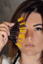 Gorgeous young lashmaker holding yellow disposable eyelash extension brushes close to face standing on gray background.