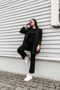 Gorgeous young woman in blazer in t-shirt in jeans in gym shoes with handbag in sunglasses is resting near vintage wooden building Royalty Free Stock Photo