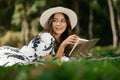 Gorgeous young woman in casual dress and straw hat reading book on picnic blanket in the park Royalty Free Stock Photo