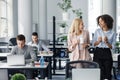 Gossip and coffee break at work. European and african american women with cups smile and talk in office interior with Royalty Free Stock Photo