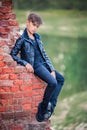 Graceful ballet boy in black leather jacket sitting on a ruined brick wall