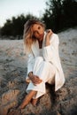 Graceful blonde in white suit sitting on sand during sunset