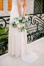 A graceful bride stands on a wrought-iron bridge in the old town of Budva and holds a bouquet behind her back, close-up