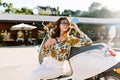 Graceful curly lady wearing white pants sitting on scooter near restaurant and mysteriously looking away. Attractive