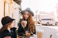 Graceful stylish woman with long light-brown hair drinking cocktail and thoughtfully looking at girl in black hat