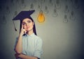 Graduate student woman in cap gown looking up at bright light bulb thinking Royalty Free Stock Photo