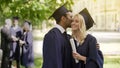 Graduates in academic regalia smiling, happy guy kissing girlfriend on cheek