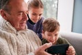 Grandchildren On Sofa At Home Showing Grandfather How To Use Mobile Phone