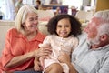 Granddaughter Cuddling With Loving Grandparents On Sofa At Home With Parents In Background