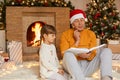 Grandfather and granddaughter reading against fireplace and xmas tree in living room, senior man holds book and looks at pages Royalty Free Stock Photo