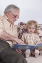 The grandfather reads a book for the grandson. The child listens carefully and looks at the book