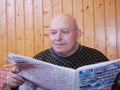 The grandfather reads the newspaper sitting on a sofa against the background of a wall from lining.