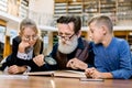 Grandfather sitting with grandchildren at the table in library, reading and telling book fairy tale story. Happy excited Royalty Free Stock Photo