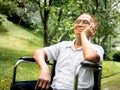 Disabled senior man in eyeglasses sitting in wheelchair looking away and thinking with his hand on chin in the park. Royalty Free Stock Photo