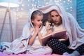 Grandmother and granddaughter are reading book under blanket at night at home. Royalty Free Stock Photo