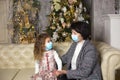 Grandmother and granddaughter on the sofa in the living room with Christmas decor hugging in medical masks on their faces. A famil