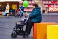 Grandmother holds an empty pram and green inflatable balloon and looks forward behind baby. Side view.