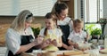 Granny with offsprings and mother in modern kitchen. Curious older daughter girl touching grandmother's apron with