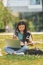 On the grass with laptop on legs. Young asian woman is outdoors at daytime