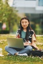 On the grass with laptop on legs. Young asian woman is outdoors at daytime