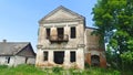 On a grassy lawn stands an old brick two-story house with a metal balcony, but no windows and doors. Next to it stand houses and g Royalty Free Stock Photo