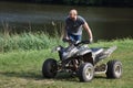 A dirty black quad and a man standing next to it. In the background is a river