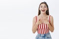 Grateful delighted young attractive woman in stylish striped top and headband holding palms on chest in thankful and