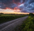 Gravel countryside road through spring meadow, cloudy evening sunset sky, rural hills, village and fields in far Royalty Free Stock Photo