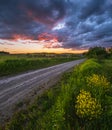 Gravel countryside road through spring meadow, cloudy evening sunset sky, rural hills, village and fields in far Royalty Free Stock Photo