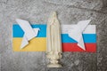 On a gray background, a figurine of the Virgin Mary, the flags of Russia and Ukraine, origami paper pigeons. Conflict between