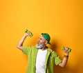 Elderly man in green cap and shirt, white t-shirt, bracelet. Smiling, doing exercises with dumbbells, posing on orange background