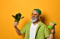 Gray-bearded grandpa in green cap and shirt, white t-shirt, sunglasses. Holding dumbbells and lettuce, posing on orange background