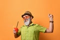 Gray-bearded man in hat, green shirt, sunglasses. Smiling, stretching himself, holding glass of juice, posing on orange background Royalty Free Stock Photo