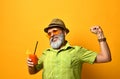 Gray-bearded man in hat, green shirt, sunglasses. Smiling, stretching himself, holding glass of juice, posing on orange background Royalty Free Stock Photo