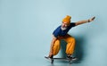 Aged man in t-shirt, orange pants, hat, gumshoes. Riding black skateboard, looking at it, posing on blue background Royalty Free Stock Photo