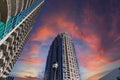 A gray and blue skyscraper with metal balconies near an office buildings with powerful clouds at sunset