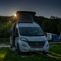 Gray camper van with a pop-up roof parked in a campground with evening light and a sun star