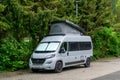 Gray camper van with a pop-up roof parked in a parking lot next to green hedges and trees