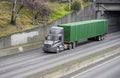 Gray day cab big rig semi truck for local deliveries transporting green container driving on the city highway road under the Royalty Free Stock Photo