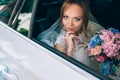 A gray-eyed blonde in a wedding dress is sitting in the car. Portrait of a pretty girl with makeup and a bouquet of Royalty Free Stock Photo