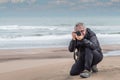 Gray haired adult man with glasses kneeling on a dune taking a photograph. Out of focus of beach and sea background.