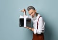 Aged male in white shirt, brown pants and suspenders, bracelet. He holding silver gift box, posing sideways on blue background Royalty Free Stock Photo