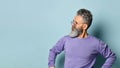 Gray-haired, bearded elderly man in sunglasses, purple sweater. Hands on hips, looking aside, posing on blue background. Close up