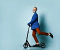 Gray-haired elderly man in white shirt, jacket, brown pants, loafers. Riding black scooter and posing sideways on blue background