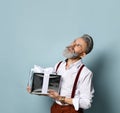 Gray-haired man in white shirt, brown pants and suspenders. Smiling, holding silver gift box, posing against blue background Royalty Free Stock Photo